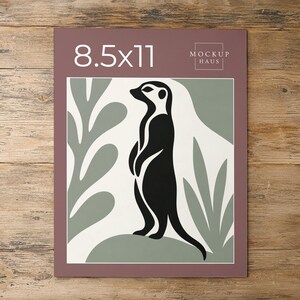 May include: A rectangular print featuring a black meerkat silhouette standing on a sage green mound against a cream background with sage green leaf shapes. The top border is mauve with the white text 8.5x11 and the logo MOCKUP HAUS.