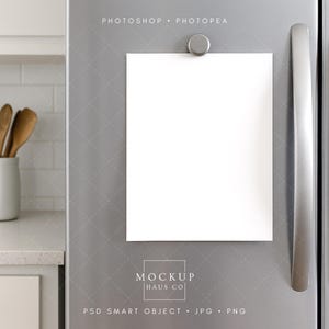 Könnte beinhalten: Ein leeres weißes Papier wird von einem silbernen Magneten an einer Edelstahl-Kühlschranktür gehalten. Das Bild ist ein Mockup mit dem Text "MOCKUP HAUS CO" und "PSD SMART OBJECT . JPG . PNG". Küchenutensilien aus Holz befinden sich in einem weißen Keramikbehälter.
