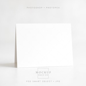 May include: A blank white greeting card stands upright on a white surface. The card is slightly angled, showcasing its front. The words "PHOTOSHOP • PHOTOPAE" are at the top, and "MOCKUP HAUS CO" and "PSD SMART OBJECT • JPG" are at the bottom.