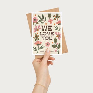 May include: A greeting card with the words "WE LOVE YOU" in brown letters, surrounded by pink and green floral illustrations. The card is held in front of a brown envelope. The words "HAPPY MOTHER'S DAY" are at the bottom.