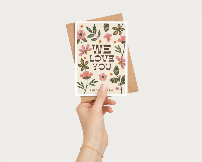 May include: A greeting card with the words "WE LOVE YOU" in brown letters, surrounded by pink and green floral illustrations. The card is held in front of a brown envelope. The words "HAPPY MOTHER'S DAY" are at the bottom.