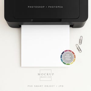 Puede incluir: Una impresora negra con una hoja de papel blanca saliendo. Dos clips plateados y una pegatina de rueda de color están junto al papel. El texto "PHOTOSHOP • PHOTOPEA" está encima del papel y "MOCKUP HAUS CO" debajo.