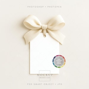 May include: A blank white gift tag with a cream-colored ribbon bow. The tag has a hole at the top and a small graphic at the bottom right. The text "ONE MOCKUP, MANY LOOKS!" is on the tag.