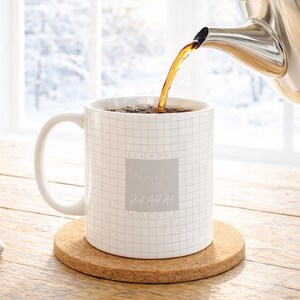 May include: A white ceramic mug with a grid pattern is being filled with tea from a stainless steel teapot. The mug sits on a round cork coaster. The text "Just All Art Mockup" is visible on the mug.