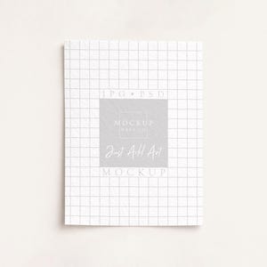 May include: A white notebook with a grid pattern, featuring the text "Just Add Art" in a soft, elegant font. The notebook is centered on a light background, creating a clean and minimalist aesthetic.