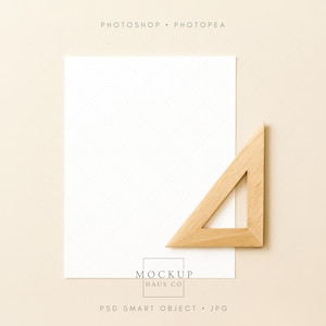 以下が含まれることがあります： ベージュ色の表面に、薄い格子模様の白い正方形の紙が置かれています。木製の三角定規が紙の上に置かれています。「PHOTOSHOP • PHOTOPAE」の文字が上部に、「MOCKUP HAUS CO」の文字が下部にあります。
