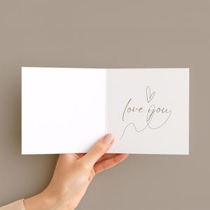May include: A white greeting card with the words "love you" written in a delicate script, accompanied by a small heart and a flowing line. The card is held open against a neutral background.