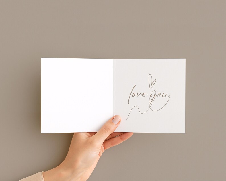 May include: A white greeting card with the words "love you" written in a delicate script, accompanied by a small heart and a flowing line. The card is held open against a neutral background.