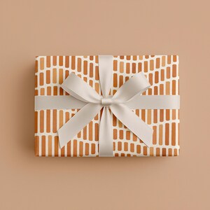 May include: A rectangular gift box wrapped in patterned paper with vertical orange and white stripes. A light gray ribbon is tied around the box with a bow on top, creating a neat and elegant presentation. The background is a solid tan color.