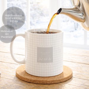 May include: A white mug with a grid pattern is filled with tea being poured from a silver teapot. The mug sits on a round cork coaster. The background includes a wooden table and a window.