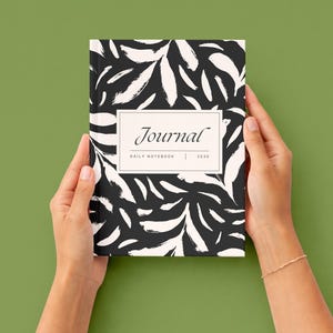 K&ouml;nnte beinhalten: Ein Notizbuch mit einem abstrakten Blattmuster in Schwarz und Wei&szlig; wird von zwei H&auml;nden gehalten. Auf dem Einband des Notizbuchs steht das Wort "Journal" in einem rechteckigen Rahmen, darunter "DAILY NOTEBOOK | 2030".