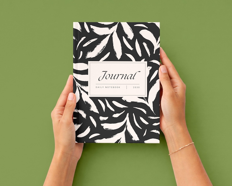 K&ouml;nnte beinhalten: Ein Notizbuch mit einem abstrakten Blattmuster in Schwarz und Wei&szlig; wird von zwei H&auml;nden gehalten. Auf dem Einband des Notizbuchs steht das Wort "Journal" in einem rechteckigen Rahmen, darunter "DAILY NOTEBOOK | 2030".