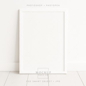 以下が含まれることがあります： 白い長方形のフォトフレームが白い壁と床に立てかけられています。 フレームは空で、上部に「PHOTOSHOP • PHOTOPEA」、下部に「MOCKUP HAUS CO」と「PSD SMART OBJECT • JPG」という文字があります。