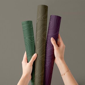 May include: Three rolled-up topographic maps in shades of green, brown, and purple. Each map features a white contour line design. The maps are held by a person against a gray background.