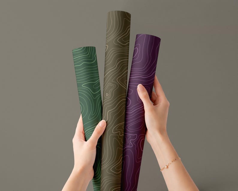 May include: Three rolled-up topographic maps in shades of green, brown, and purple. Each map features a white contour line design. The maps are held by a person against a gray background.