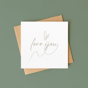 以下が含まれることがあります： 繊細な筆記体で「love you」と書かれた白い正方形のカード。小さなハートが添えられています。茶色の封筒の上に置かれ、くすんだ緑色の背景に配置されています。シンプルでエレガントなデザインです。