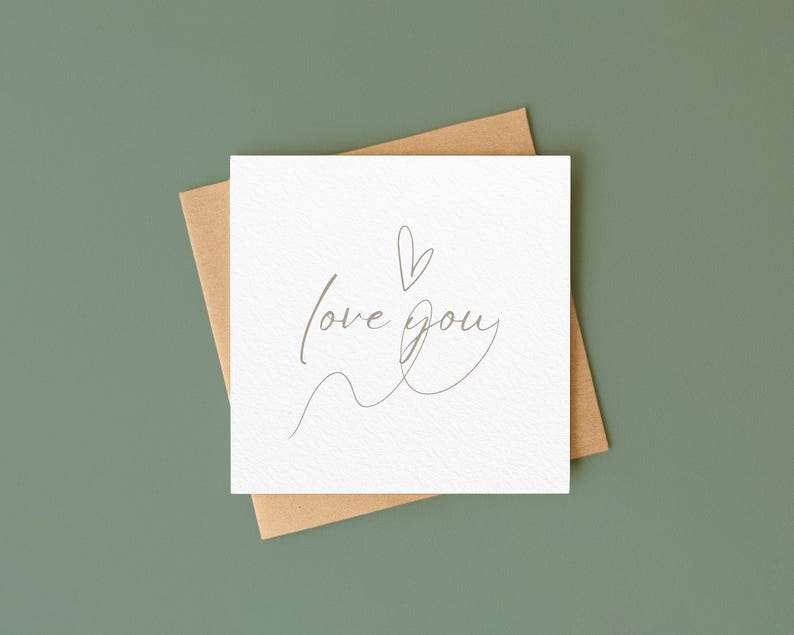 以下が含まれることがあります： 繊細な筆記体で「love you」と書かれた白い正方形のカード。小さなハートが添えられています。茶色の封筒の上に置かれ、くすんだ緑色の背景に配置されています。シンプルでエレガントなデザインです。