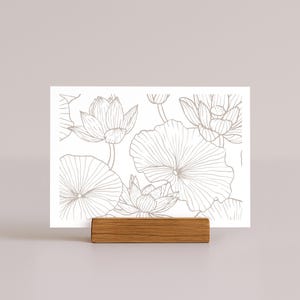 May include: A white card featuring a detailed line drawing of lotus flowers and leaves in a neutral color. The card is held by a small, rectangular wooden block. The design is suitable for stationery or art prints.