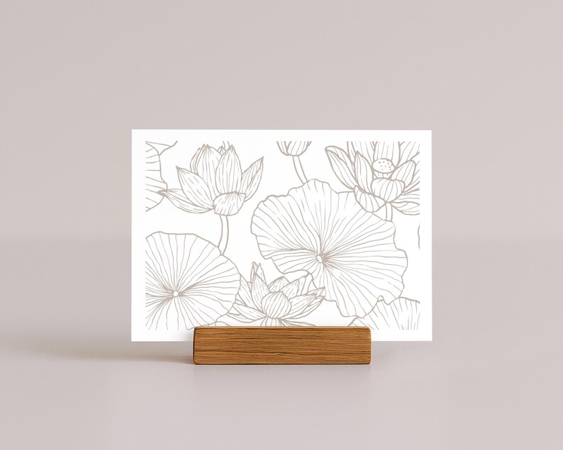 May include: A white card featuring a detailed line drawing of lotus flowers and leaves in a neutral color. The card is held by a small, rectangular wooden block. The design is suitable for stationery or art prints.