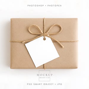 Pode incluir: Uma embalagem retangular embrulhada em papel castanho, amarrada com barbante e adornada com um laço de barbante. Uma etiqueta branca em branco pende do laço. O texto "PHOTOSHOP • PHOTOPEA" e "MOCKUP HAUS CO" está impresso.