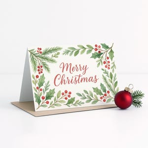 May include: A folded Christmas card with the words "Merry Christmas" in red script, surrounded by a watercolor design of green leaves and red berries. A red Christmas ornament and sprig of pine are next to the card.