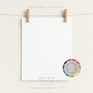 Op de afbeelding: Een wit, getextureerd vel papier wordt vastgehouden door twee houten wasknijpers aan een dun touwtje tegen een crèmekleurige achtergrond. Het papier heeft de tekst "MOCKUP HAUS CO" en "PSD SMART OBJECT • JPG". Een kleurenwiel-afbeelding is ook aanwezig.