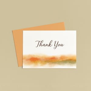 K&ouml;nnte beinhalten: Eine wei&szlig;e Karte mit dem Schriftzug "Thank You" in eleganter Schrift, &uuml;ber einer Aquarelllandschaft in Orange-, Gelb- und Gr&uuml;nt&ouml;nen. Ein orangefarbener Umschlag liegt hinter der Karte, vor einem neutralen Hintergrund.