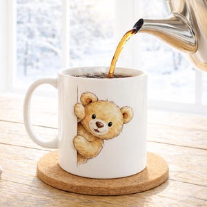 May include: A white ceramic mug featuring a cartoon teddy bear design, with a brown cork coaster underneath. Hot liquid is being poured from a stainless steel teapot into the mug. The background shows a snowy winter scene.