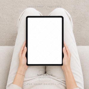 May include: A black tablet with a blank white screen is held by a person wearing a beige sweater and pants. The tablet is centered, with the person's hands visible. The background is a neutral beige.