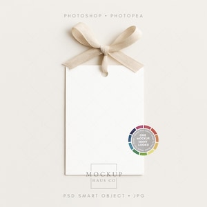 Pode incluir: Uma etiqueta retangular branca em branco com um laço de fita bege na parte superior. A etiqueta tem um pequeno orifício para pendurar. Uma roda de cores com o texto "ONE MOCKUP MANY LOOKS!" está à direita. O texto "MOCKUP HAUS CO" está na parte inferior.