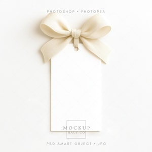 Puede incluir: Una etiqueta de regalo blanca en blanco con un lazo color crema en la parte superior. La etiqueta tiene un pequeño agujero para sujetarla a un regalo. El texto "PHOTOSHOP • PHOTOPEA" está en la parte superior, y "MOCKUP HAUS CO" y "PSD SMART OBJECT • JPG" están en la parte inferior.