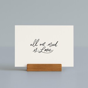 May include: A cream-colored card with the handwritten phrase "all we need is Love" in black script. The card is displayed in a small, rectangular wooden holder. The background is a solid, light gray color.