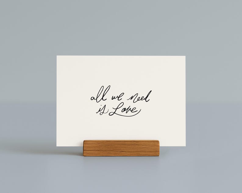 May include: A cream-colored card with the handwritten phrase "all we need is Love" in black script. The card is displayed in a small, rectangular wooden holder. The background is a solid, light gray color.