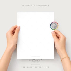 Puede incluir: Un papel blanco sostenido por dos manos, con una pegatina de rueda de color en la esquina superior derecha. El papel tiene un sutil patr&oacute;n de diamantes. El texto "MOCKUP HAUS CO" y "PSD SMART OBJECT . JPG" son visibles.