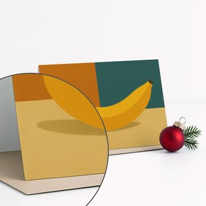 May include: Greeting card featuring a yellow banana illustration against a backdrop of mustard yellow, teal, and orange blocks. A red Christmas ornament with a small sprig of greenery sits to the right of the card. The card is displayed on a white surface.