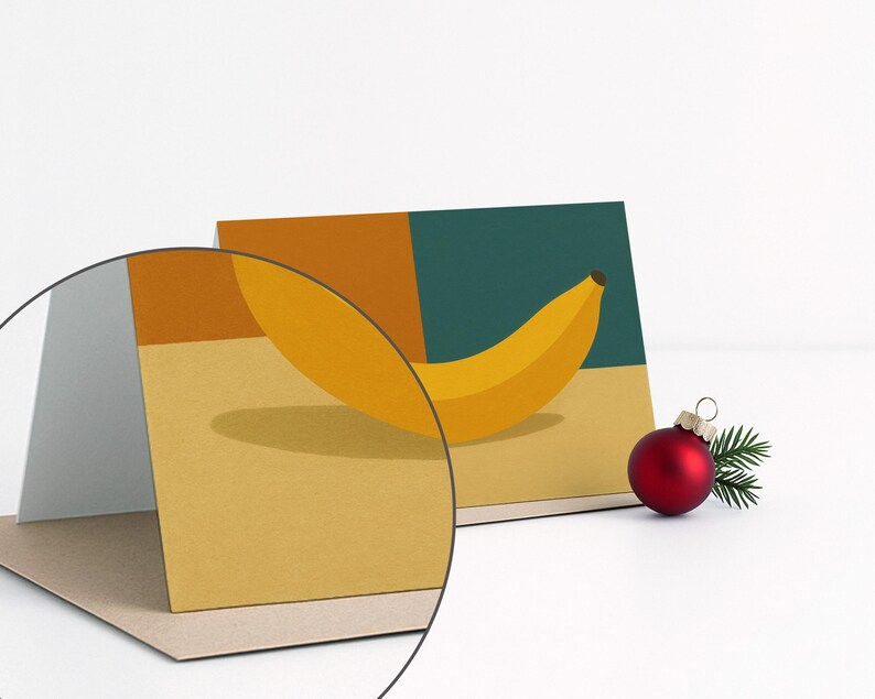 May include: Greeting card featuring a yellow banana illustration against a backdrop of mustard yellow, teal, and orange blocks. A red Christmas ornament with a small sprig of greenery sits to the right of the card. The card is displayed on a white surface.