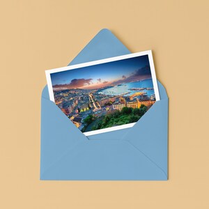 May include: A postcard featuring a scenic city view, partially visible within a light blue envelope. The postcard showcases a coastal cityscape with buildings, roads, and a body of water under a colorful sunset sky. The envelope is on a tan surface.