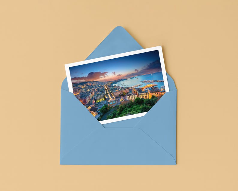 May include: A postcard featuring a scenic city view, partially visible within a light blue envelope. The postcard showcases a coastal cityscape with buildings, roads, and a body of water under a colorful sunset sky. The envelope is on a tan surface.