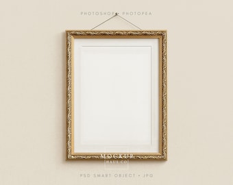 Op de afbeelding: Een rechthoekige gouden fotolijst met sierlijke details hangt aan een crèmekleurige muur. De lijst heeft een witte passe-partout en een lege witte ruimte voor een afbeelding. De tekst "PHOTOSHOP PHOTOPAE" staat bovenaan en "MOCKUR HAUS CO" en "PSD SMART OBJECT . JPG" staan onderaan.