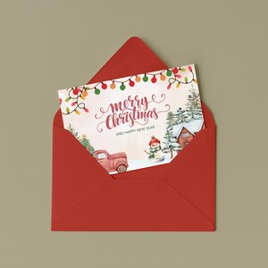 May include: A red envelope containing a Christmas card. The card features a watercolor illustration of a red truck, snowman, and a house in a snowy landscape. The card reads "Merry Christmas and Happy New Year" with festive string lights.