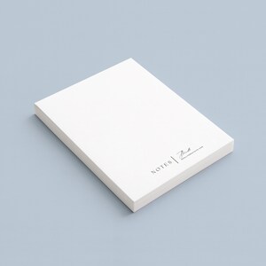 Peut inclure: Un bloc-notes blanc rectangulaire sur une surface bleu clair. Le papier a un design minimaliste. Le coin inf&eacute;rieur droit du papier porte le mot "NOTES" et une signature en noir. Le fond est bleu clair uni.