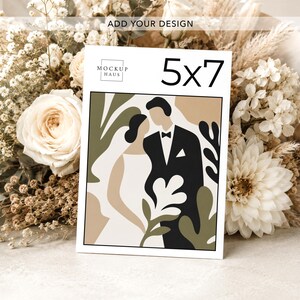 Pu&ograve; includere: Un biglietto di nozze di 12,7 x 17,8 cm con un'illustrazione astratta di una sposa e uno sposo, circondati da forme verde oliva e beige. Il biglietto &egrave; esposto davanti a un bouquet di fiori bianchi e beige.
