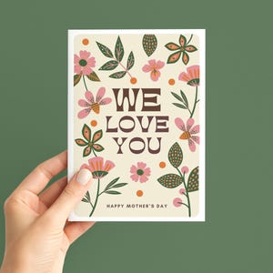 May include: A cream-coloured greeting card featuring the text "WE LOVE YOU" in brown, surrounded by pink and orange floral illustrations and green leaves. The words "HAPPY MOTHER'S DAY" are at the bottom.