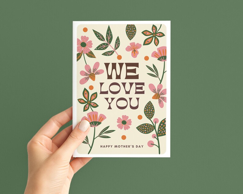 May include: A cream-coloured greeting card featuring the text "WE LOVE YOU" in brown, surrounded by pink and orange floral illustrations and green leaves. The words "HAPPY MOTHER'S DAY" are at the bottom.