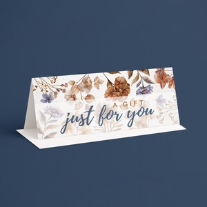 Pu&ograve; includere: Un biglietto regalo bianco con un design floreale con fiori marroni, blu e bianchi. Il biglietto recita "A Gift Just For You" in un carattere corsivo. Il biglietto &egrave; piegato e si erge su una base bianca su uno sfondo blu scuro.