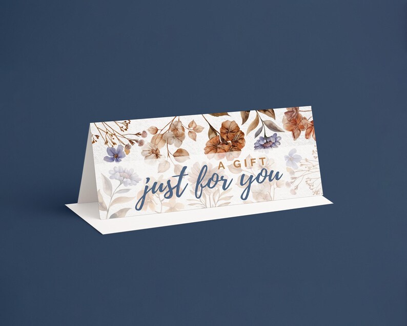 Pu&ograve; includere: Un biglietto regalo bianco con un design floreale con fiori marroni, blu e bianchi. Il biglietto recita "A Gift Just For You" in un carattere corsivo. Il biglietto &egrave; piegato e si erge su una base bianca su uno sfondo blu scuro.