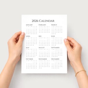 Puede incluir: Un calendario blanco de 2026 con texto negro es sostenido por dos manos. El calendario muestra los meses del a&ntilde;o en formato de cuadr&iacute;cula. El texto "2026 CALENDAR" est&aacute; en la parte superior.