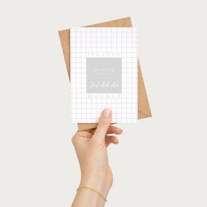 May include: A white grid-patterned card with the text "MOCKUP" and "Just All Art" is held in front of a brown envelope. The card is held by a person. The background is a solid, neutral color.