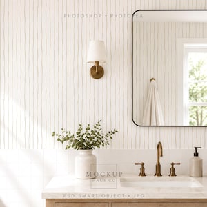 Puede incluir: Un mueble de baño con encimera blanca, grifo de latón, dispensador de jabón y un jarrón blanco con follaje verde. Una lámpara de pared y un espejo rectangular con marco negro están montados en una pared con papel tapiz de rayas verticales.