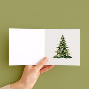 May include: A blank greeting card held by a hand, featuring a watercolor illustration of a green Christmas tree with white accents. The card is open, with the tree on the right side against a white background.